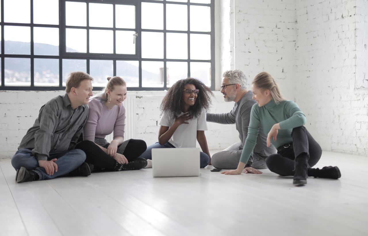 Happy multiethnic group of people gathering together while speaking and sitting with crossed legs in front of portable computer on ceramic floor near window in sunlight